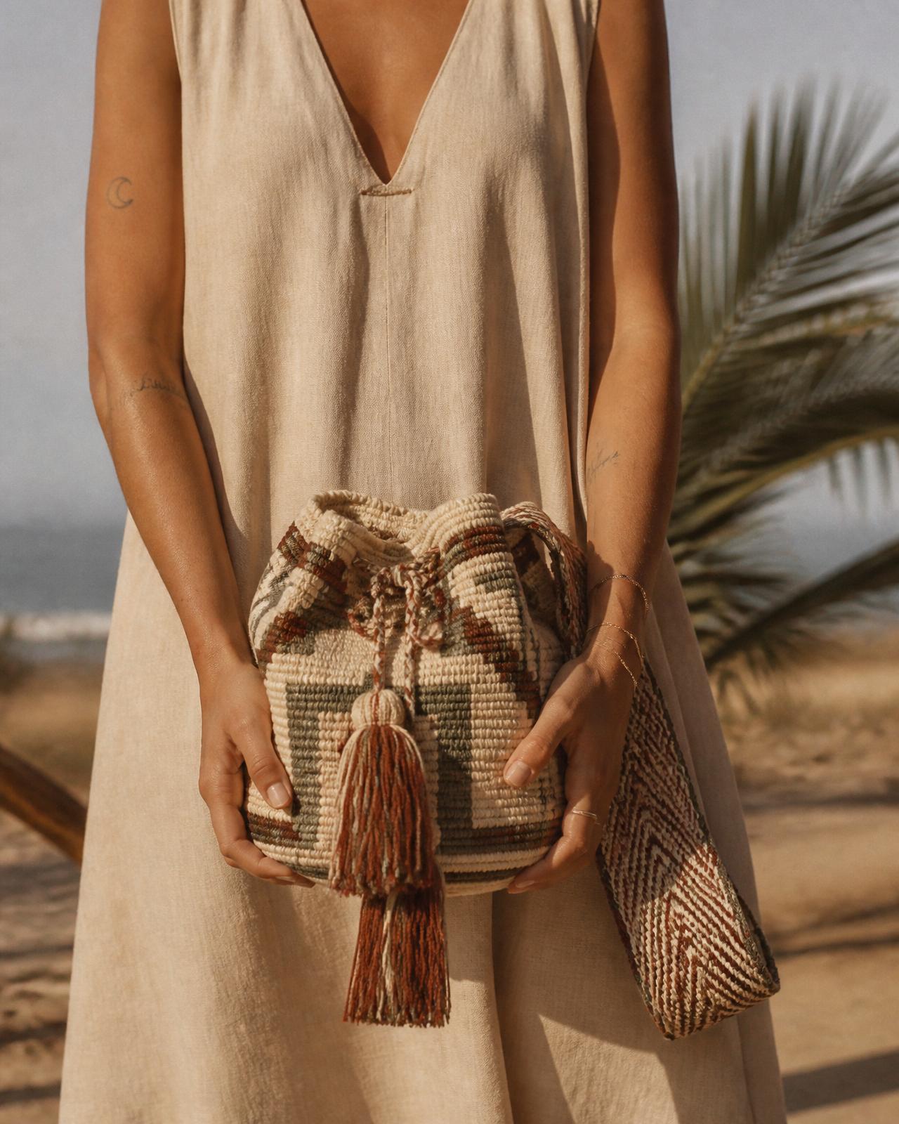 Close-up studio photograph of a woman in a linen dress holding two handwoven Wayuu mochilas at the beach